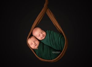 Twin newborns swaddled in green wrap, nestled together in a teardrop-shaped brown fabric holder on black background.