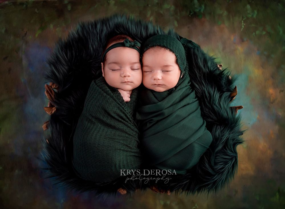Twin newborns swaddled in matching green wraps, sleeping side by side in a dark fur bowl on colorful background.