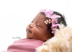 Sleeping newborn baby girl wearing a pink floral headband, wrapped in a pink swaddle, smiling peacefully.