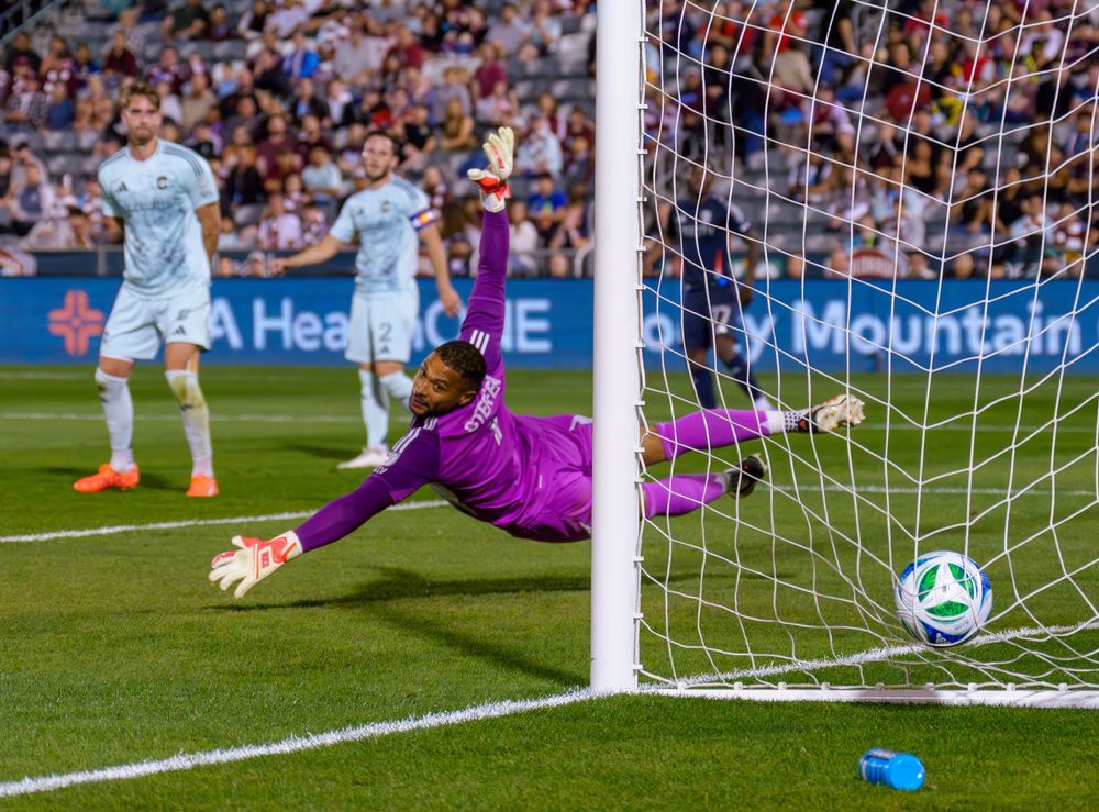 Soccer player scores goal with ball crossing goal line as goalkeeper dives to make save during match.