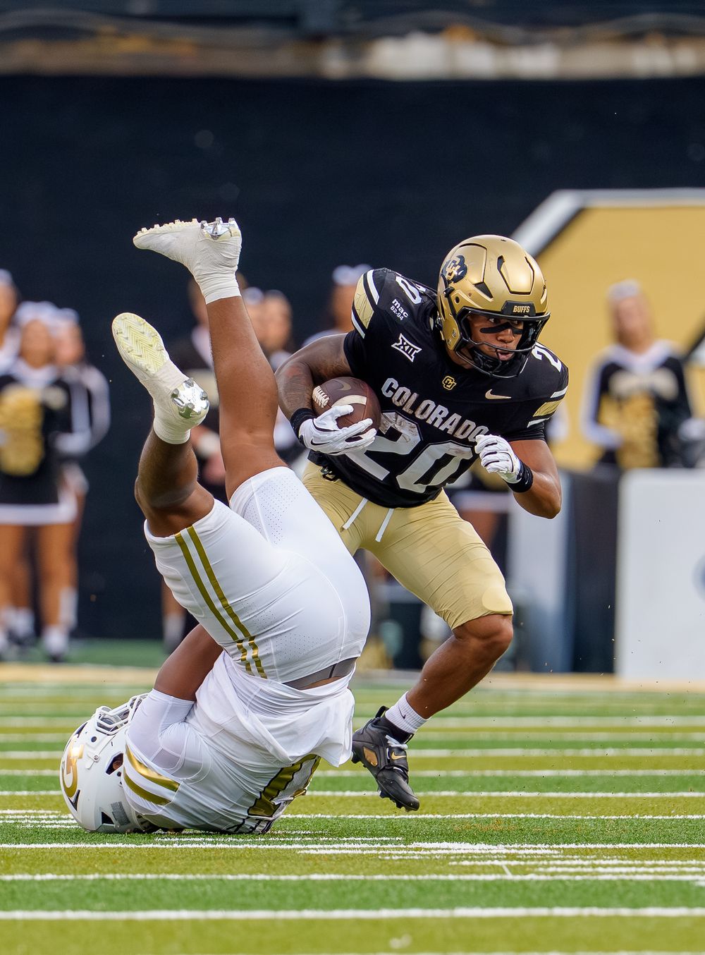 A football player in black and gold uniform falls forward while being tackled during a game on the field.