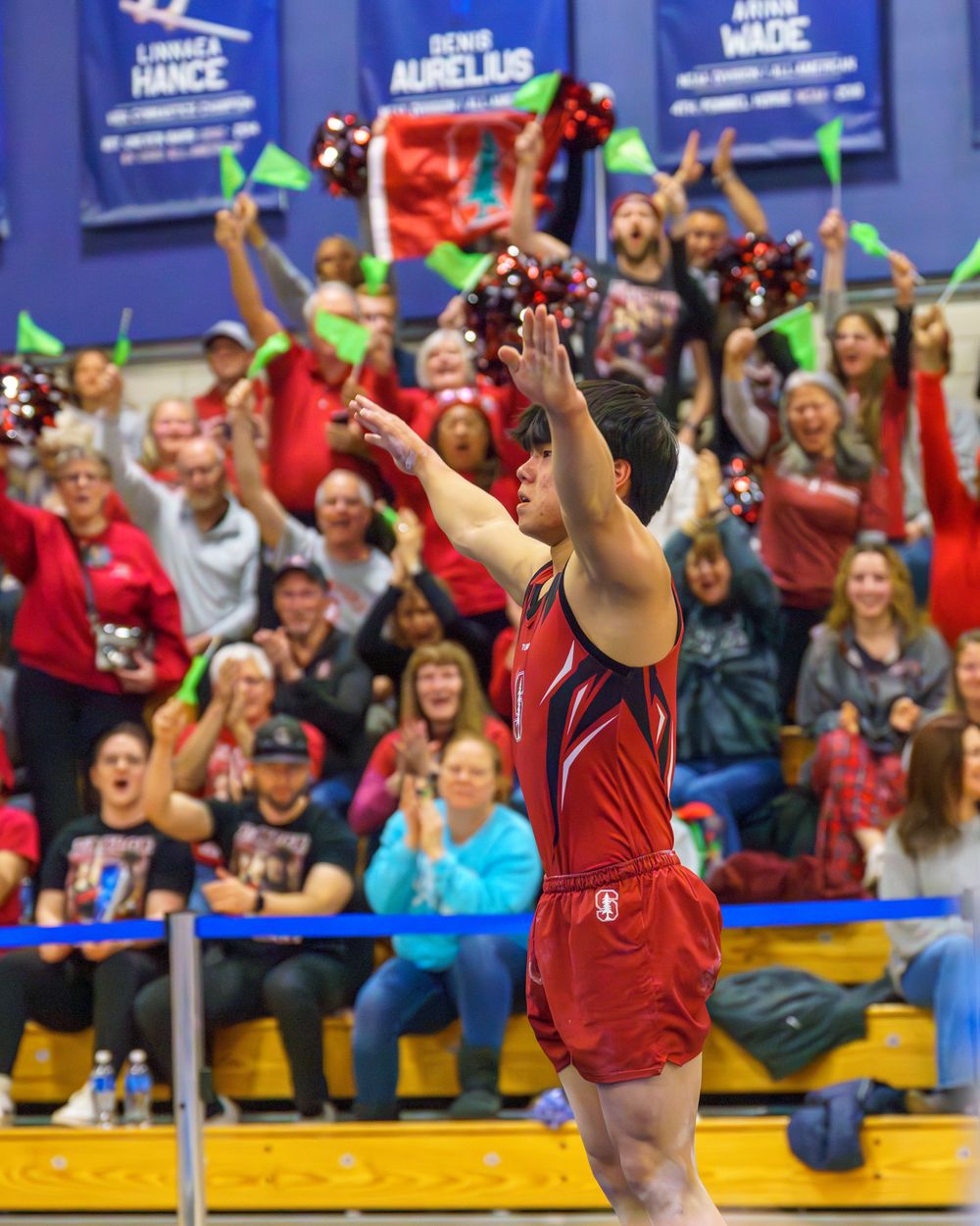 Gymnast in red performs an aerial routine with crowd cheering in background at indoor competition.