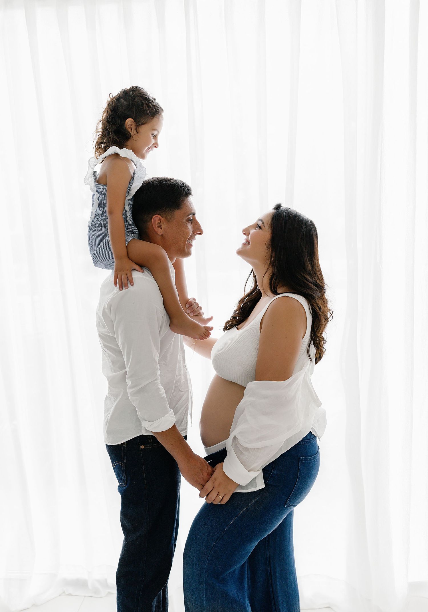 Family wearing matching white tops and blue jeans pose together against white curtain background.
