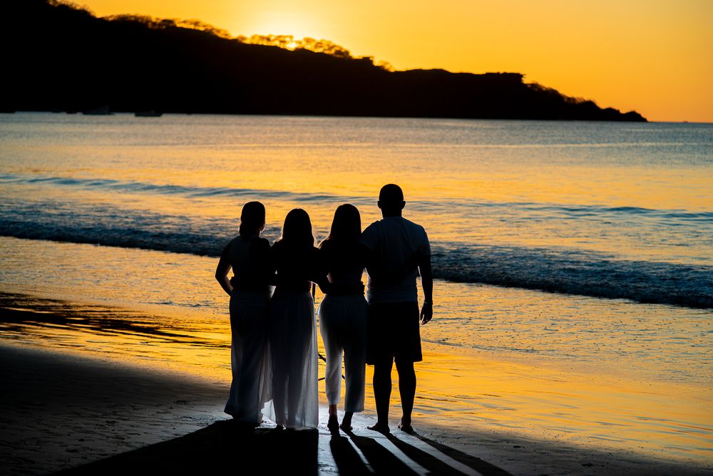 Silhouettes of people standing together on a beach at sunset with golden sky and ocean waves in the background.