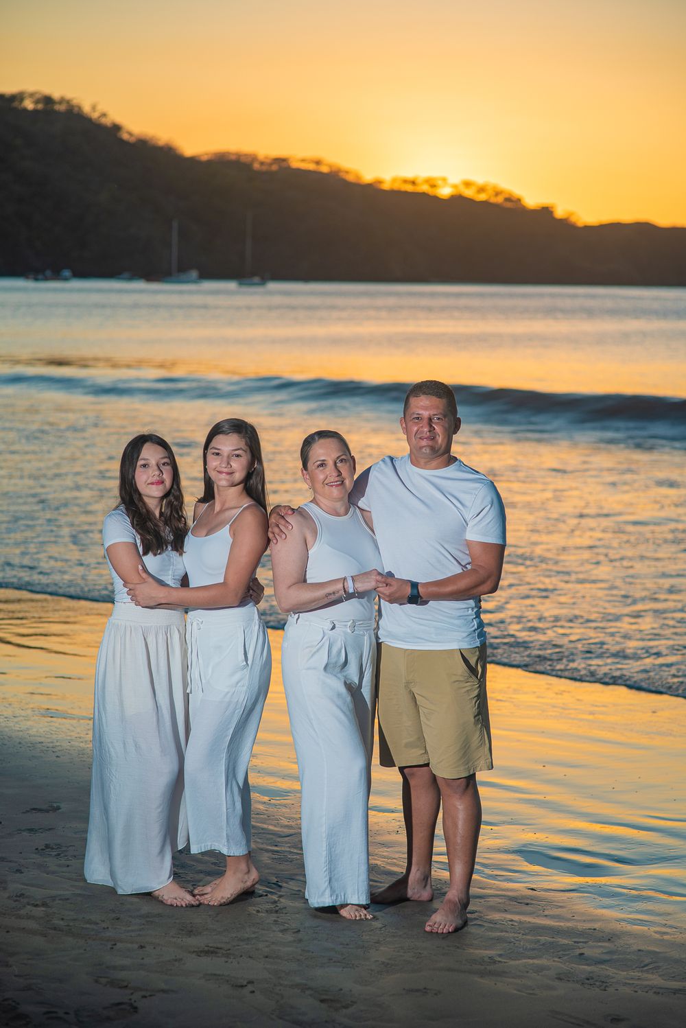 A group in white attire poses together on a beach at sunset with golden light reflecting on the water.