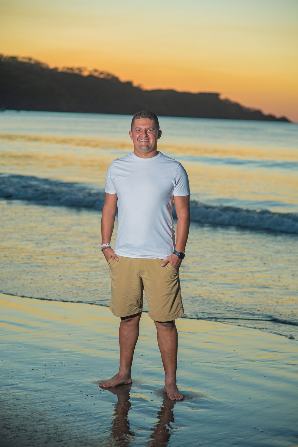 A person stands on a sandy beach at sunset with golden light reflecting off the ocean waves in the background.
