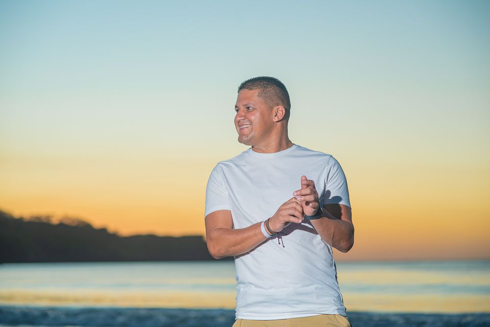 Person in white t-shirt stands against sunset sky at beach while taking photos with vintage camera.