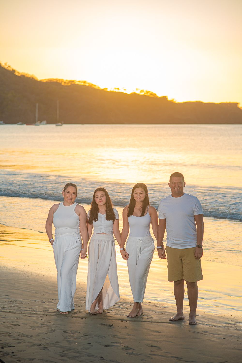 Family photoshoot in Guanacaste in white dresses and casual attire poses on a beach at sunset with golden light illuminating the coastline.