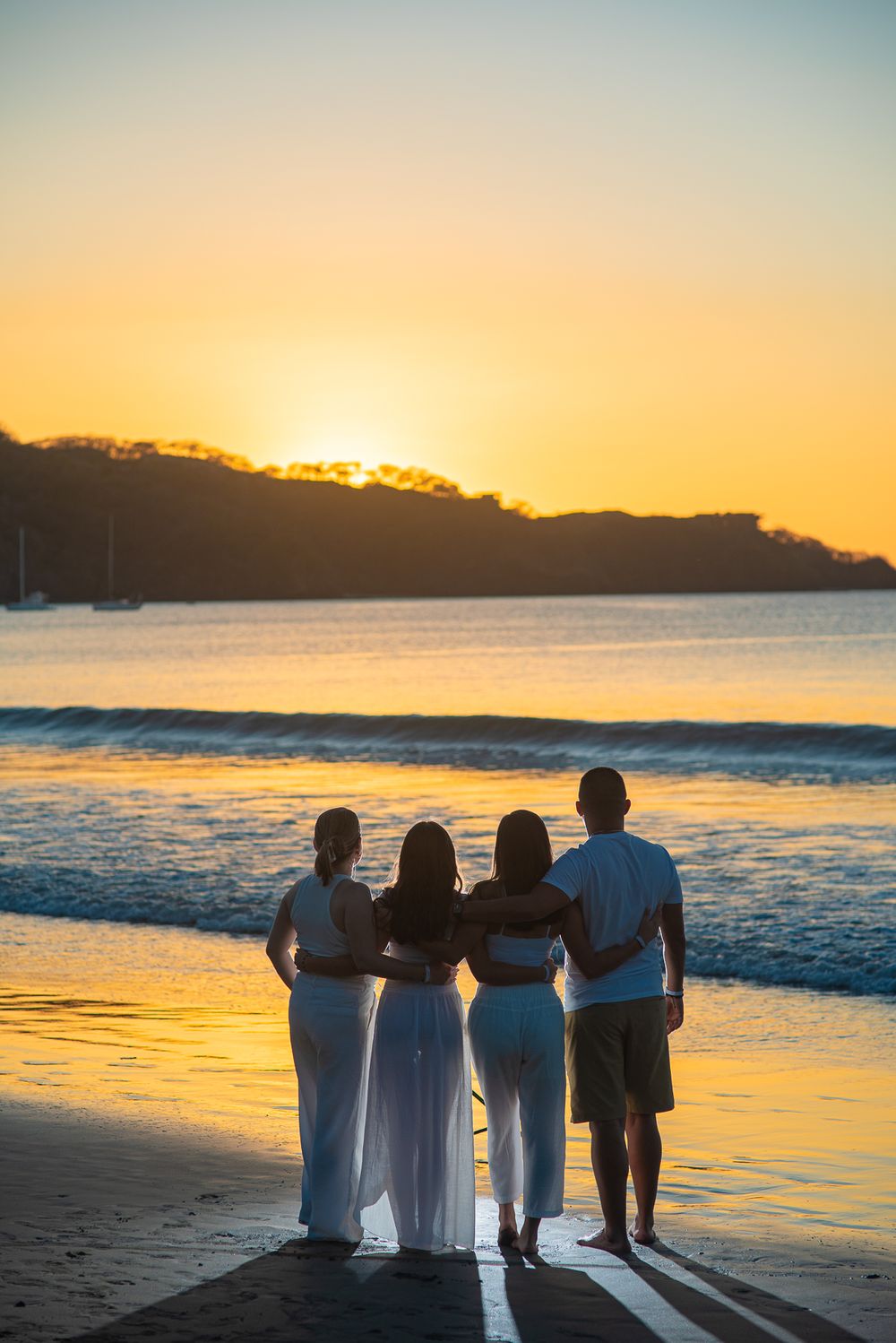 Family photoshoot in Guanacaste silhouetted figures stand together on beach watching golden sunset over ocean waves.