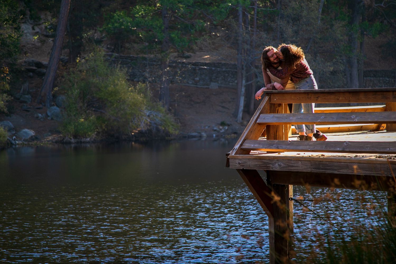 A man and woman lean against a tree outdoors, smiling at each other in warm sunlight near a pond.