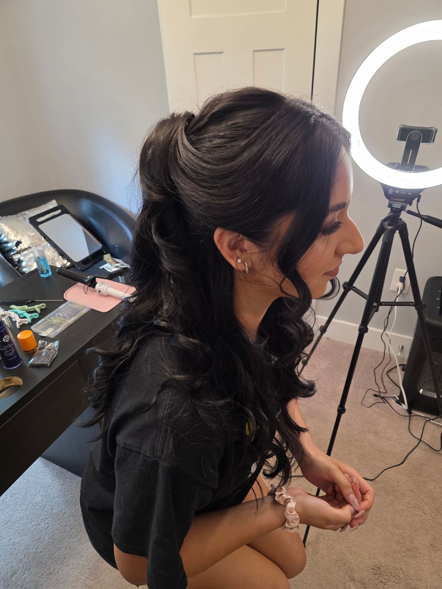 Woman with dark curly half-up hairstyle sits near a ring light and vanity mirror in a beauty studio.