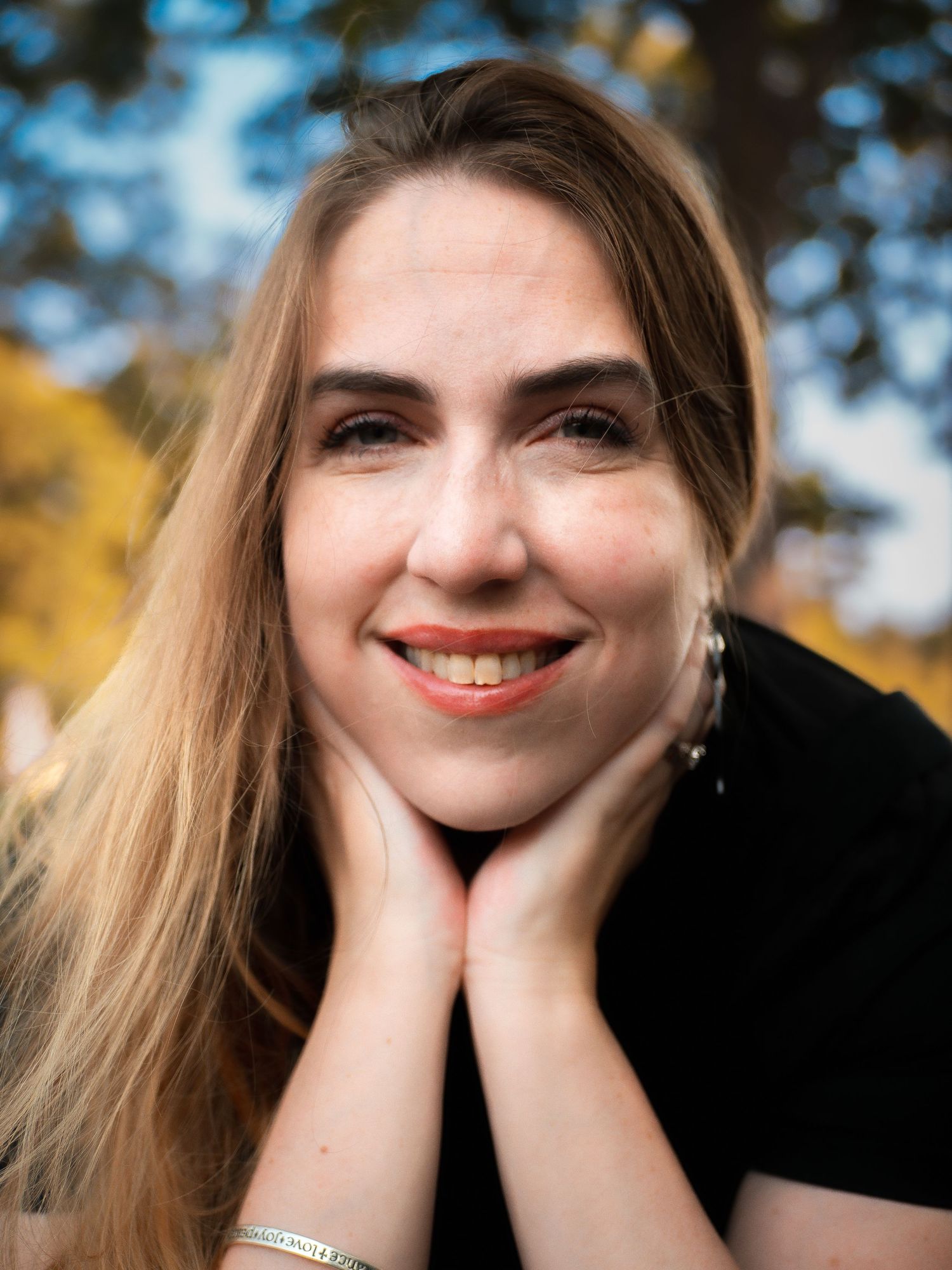 Portrait shot outdoors with autumn colors in background showing a warm smile and hands framing face.