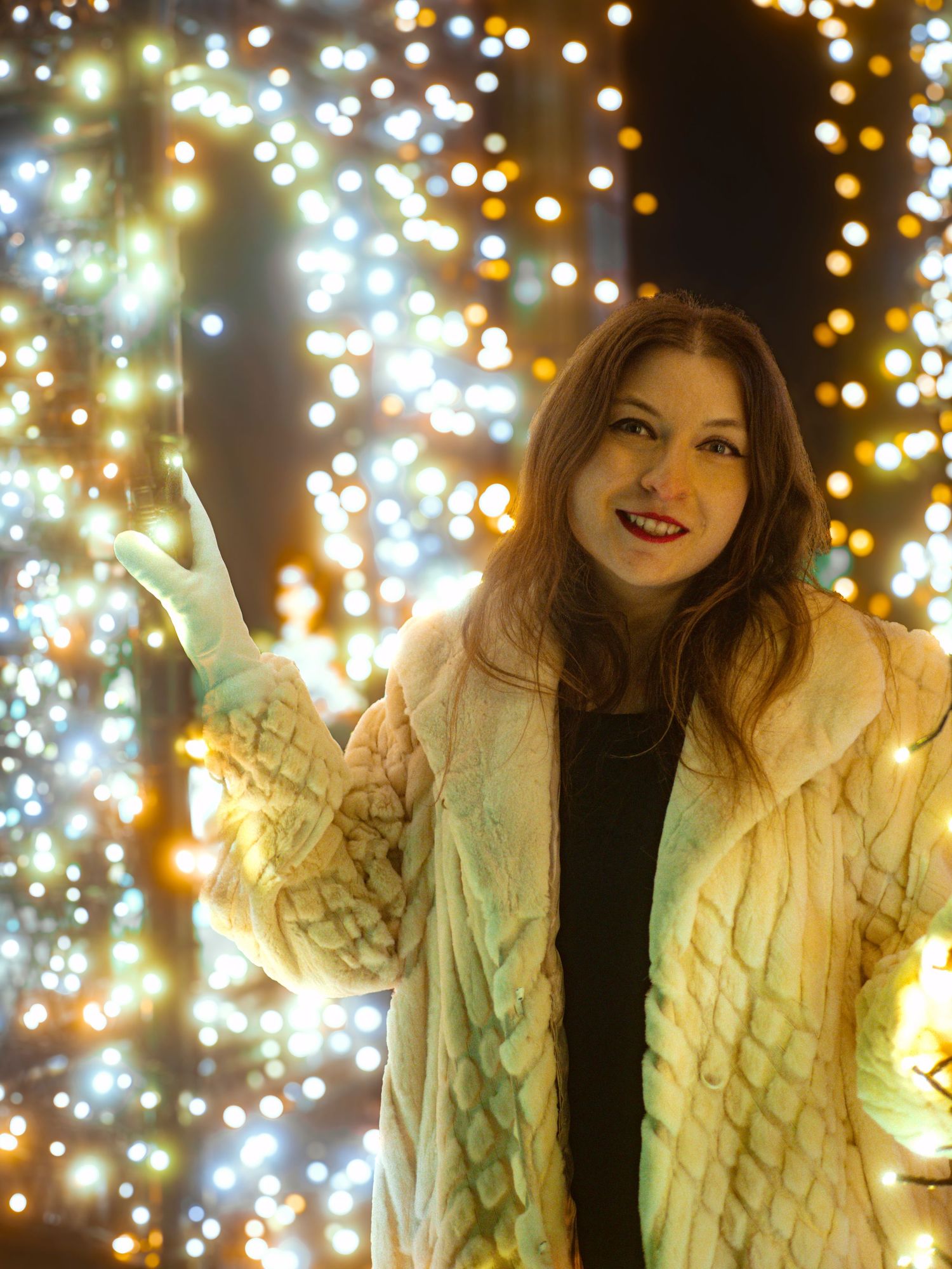 Person in white knit sweater poses against backdrop of warm glowing string lights.