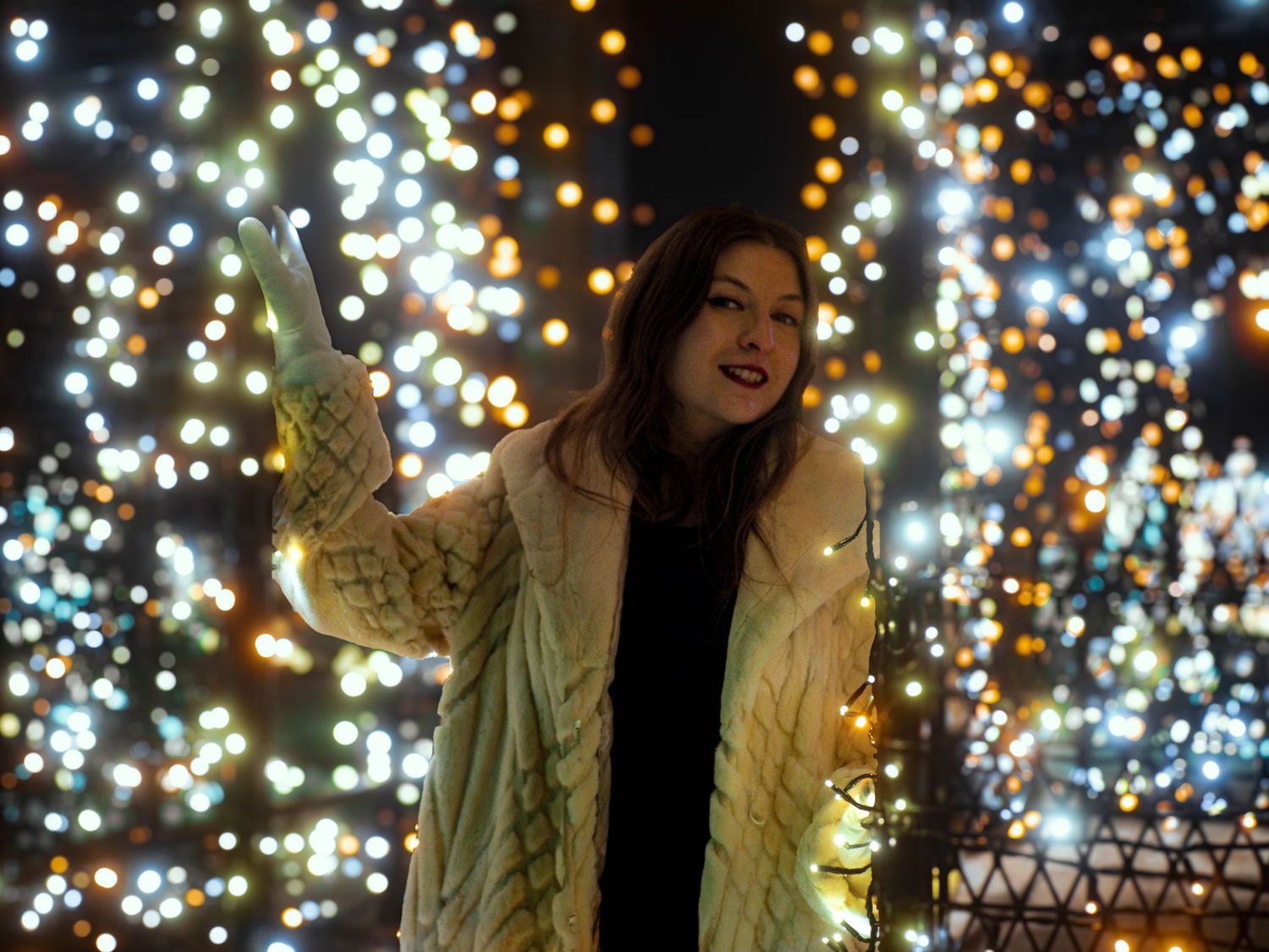 A person stands among twinkling holiday lights creating a magical bokeh effect in the nighttime scene.