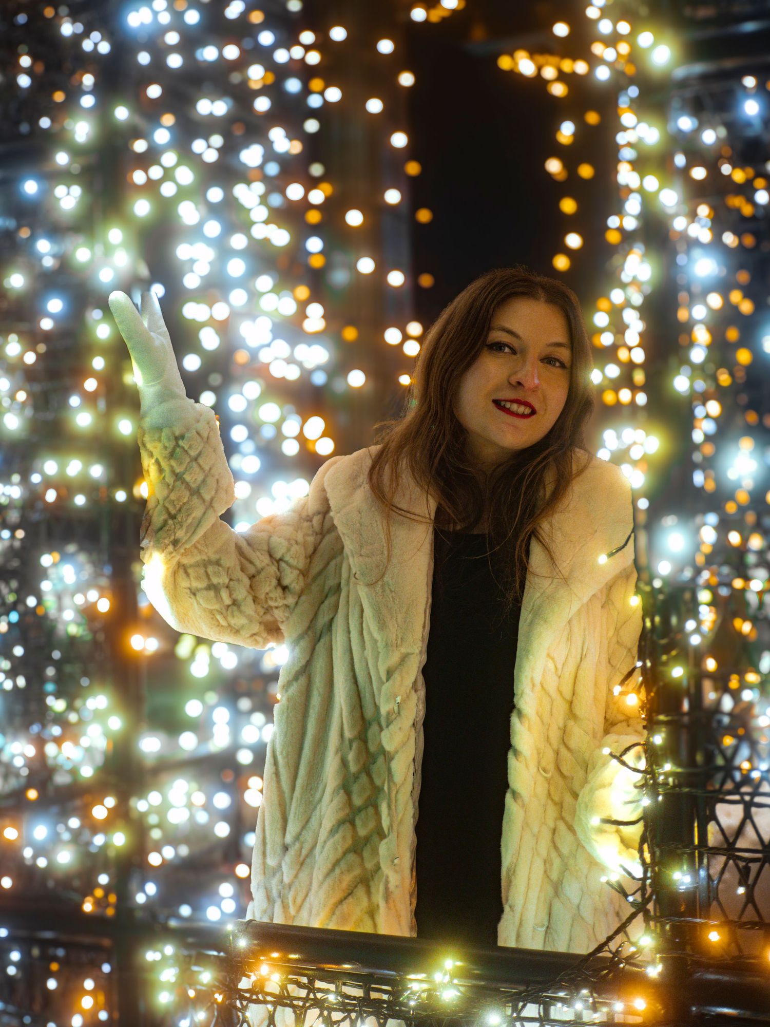 Figure in white jacket poses against sparkling holiday light backdrop.