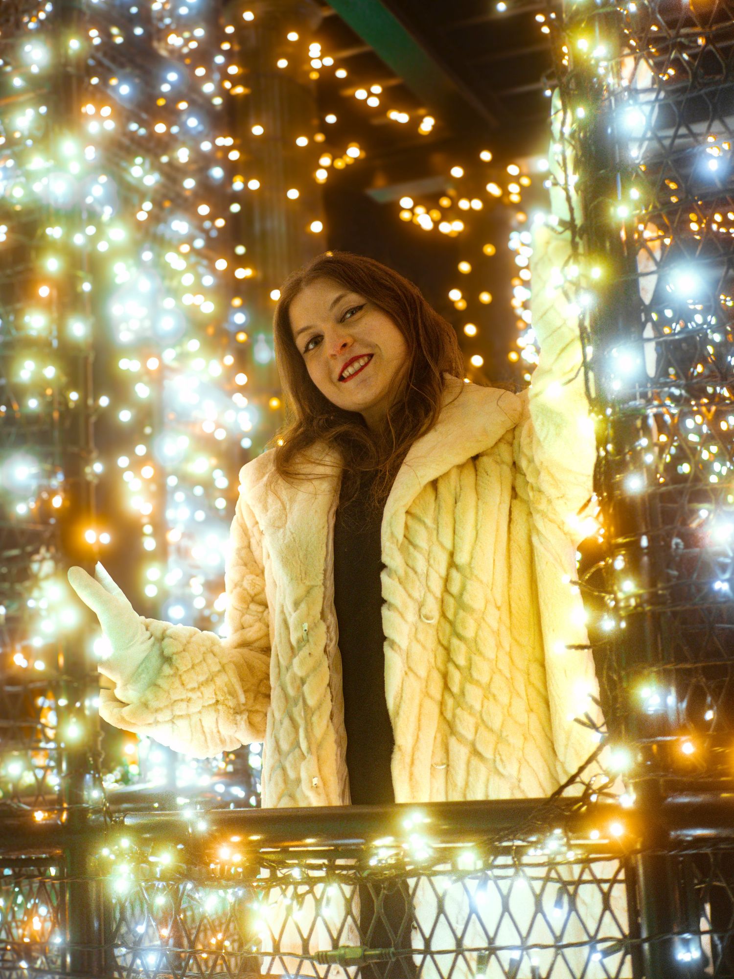 A person in a cream quilted jacket poses among twinkling holiday lights creating a magical nighttime ambiance.