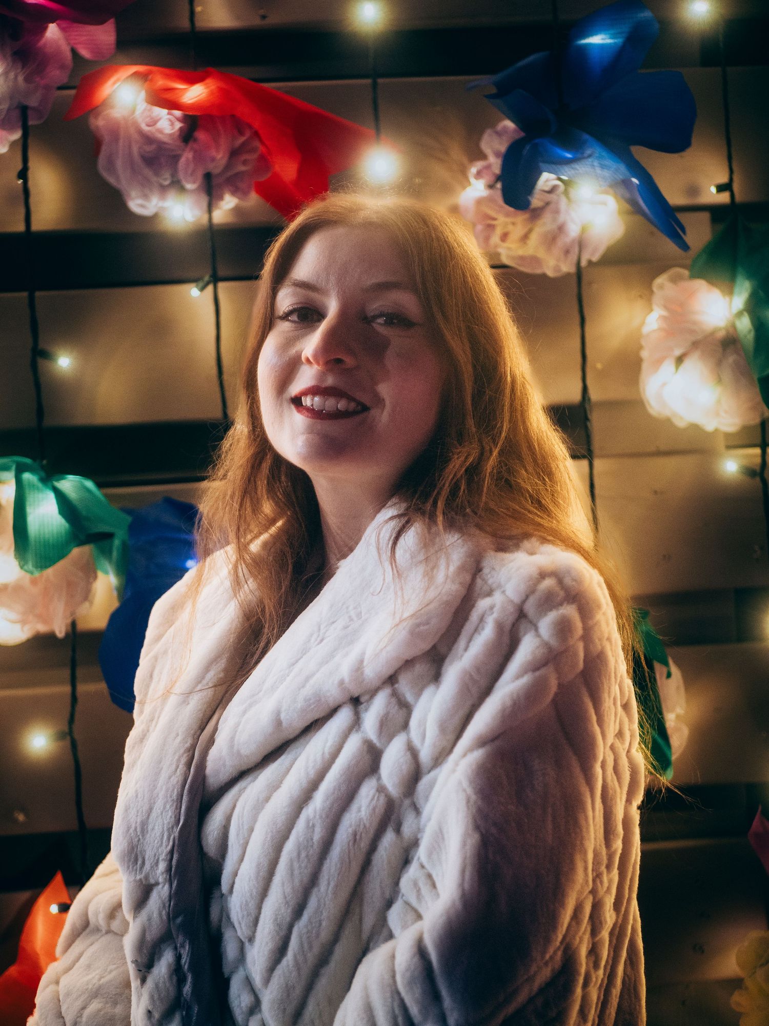 A person in a white fur coat poses under decorative string lights with colorful flowers in the background.