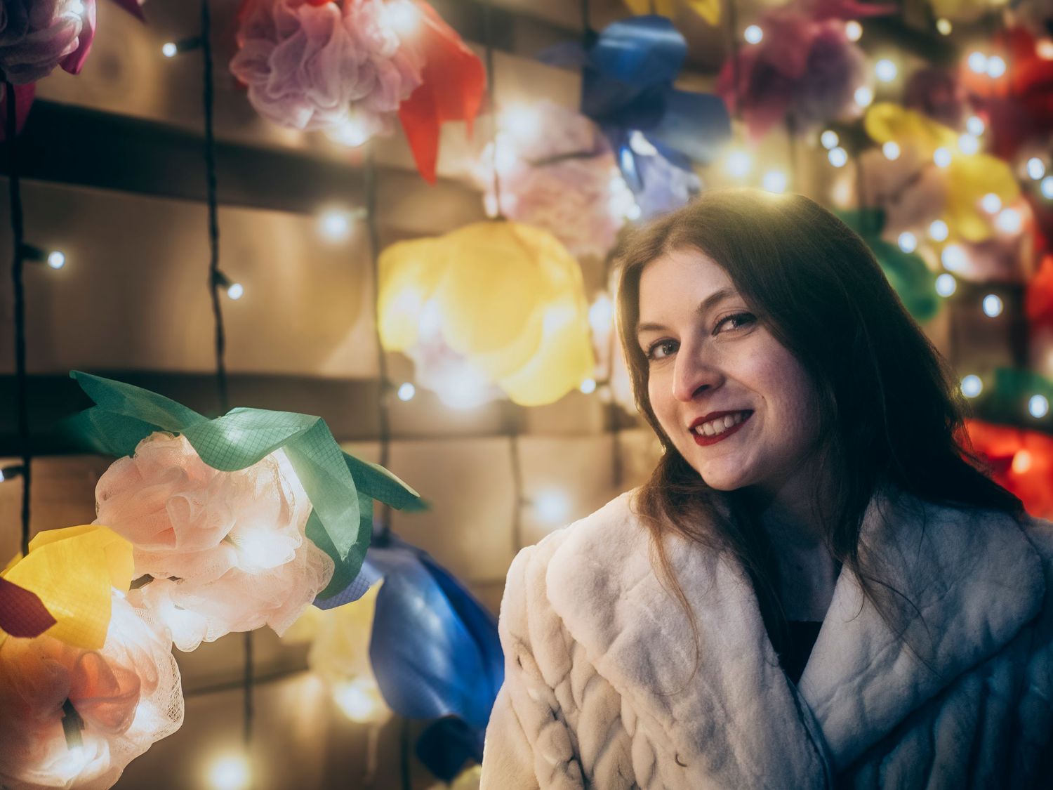 A person in a cozy white sweater smiles warmly amid colorful Christmas lights and decorative paper lanterns.