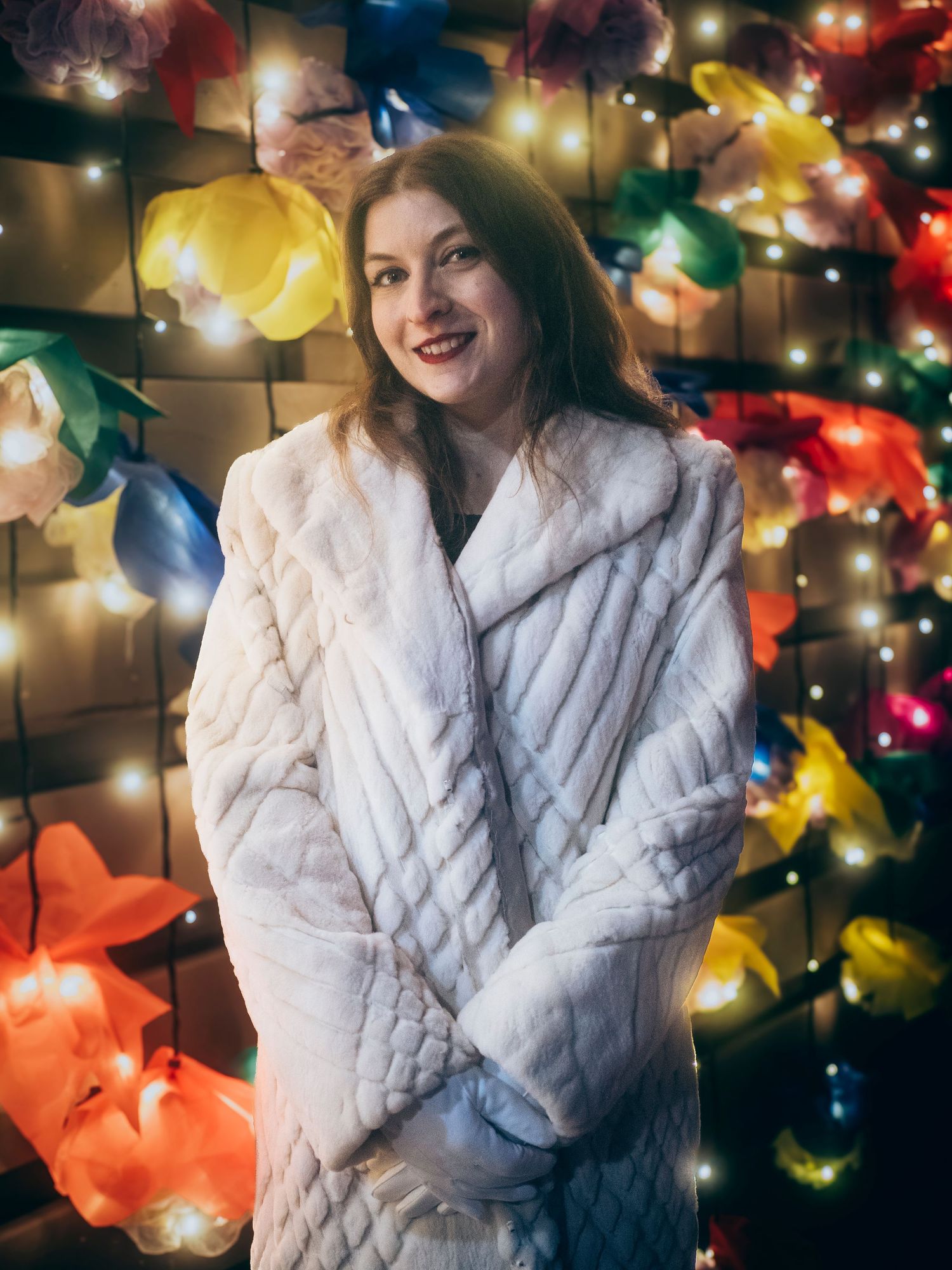Person in white quilted jacket poses among colorful holiday lights and decorations.