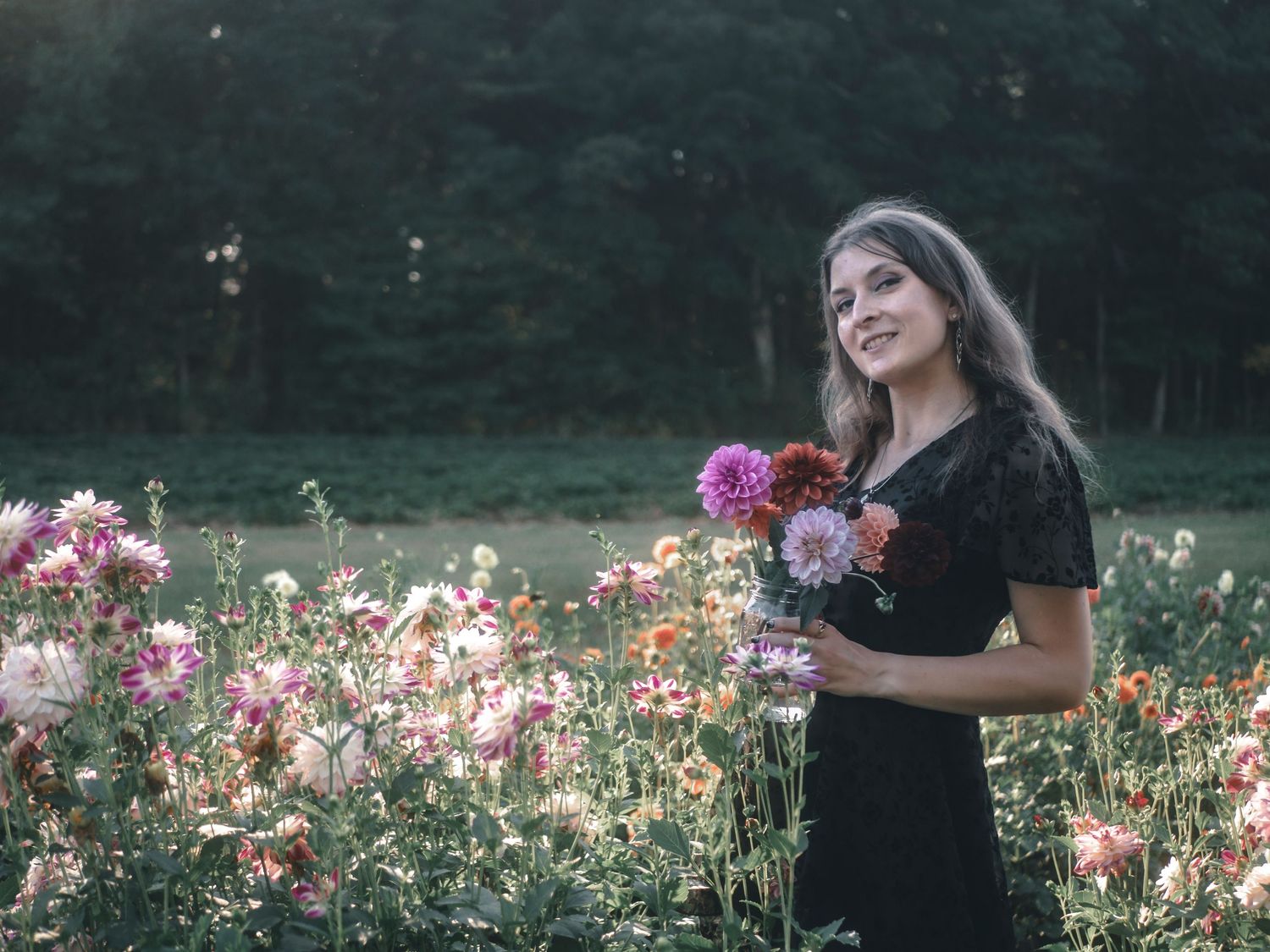 A person in a black dress holds a bouquet of colorful wildflowers in a sunlit meadow filled with blooms.