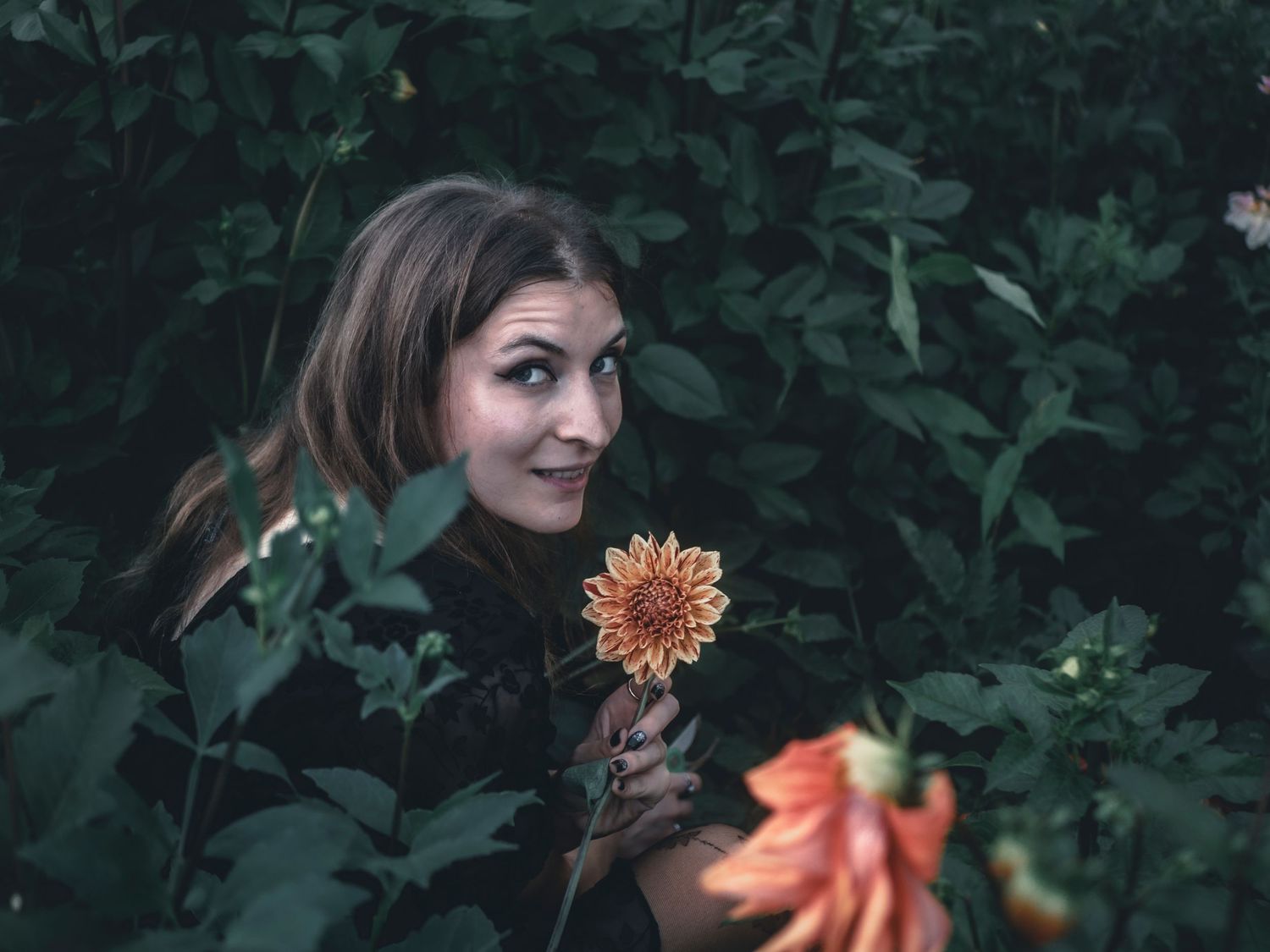 Woman in dark garden setting poses with vibrant orange dahlia flowers against lush green foliage background.