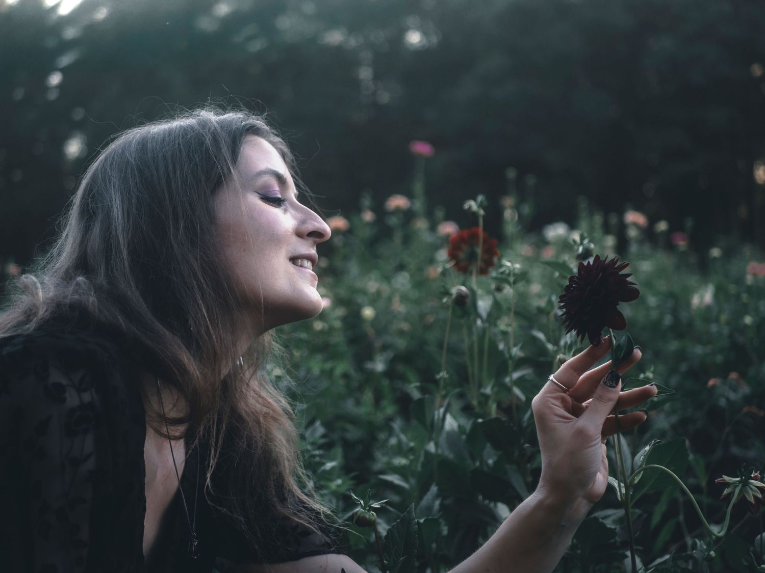 A person smiles peacefully while holding a dark flower in a dreamy garden setting at dusk.