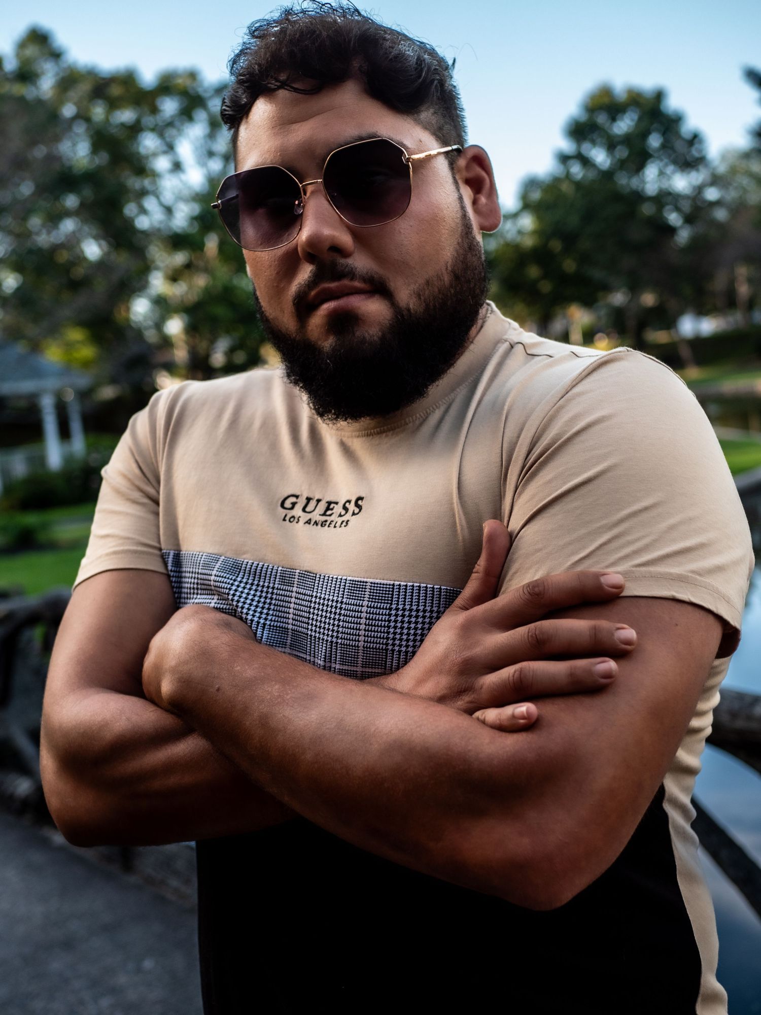 Person wearing sunglasses and beige t-shirt poses with crossed arms against outdoor background.