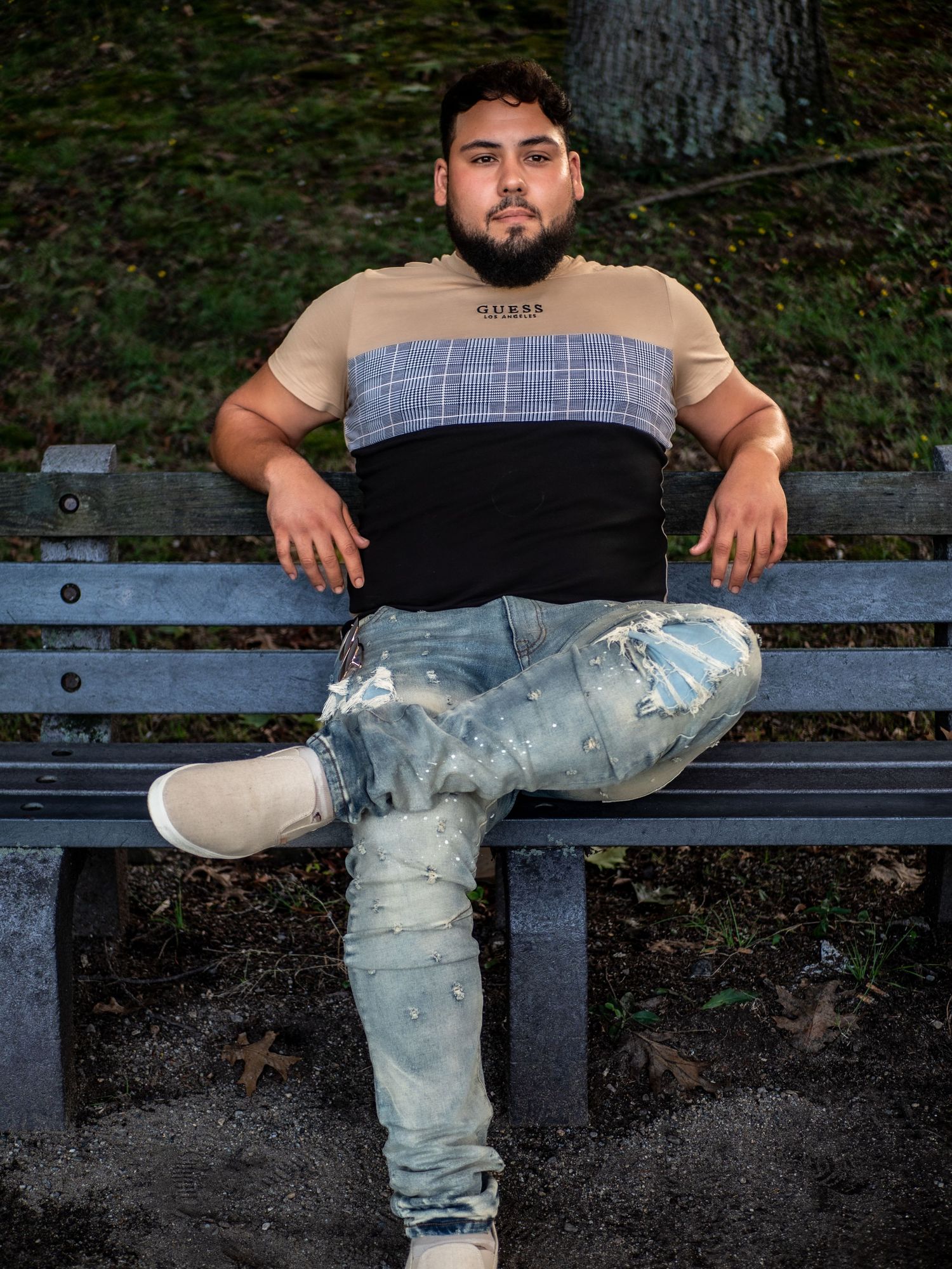 A person in ripped light-colored jeans sits casually on a dark park bench in an outdoor setting.