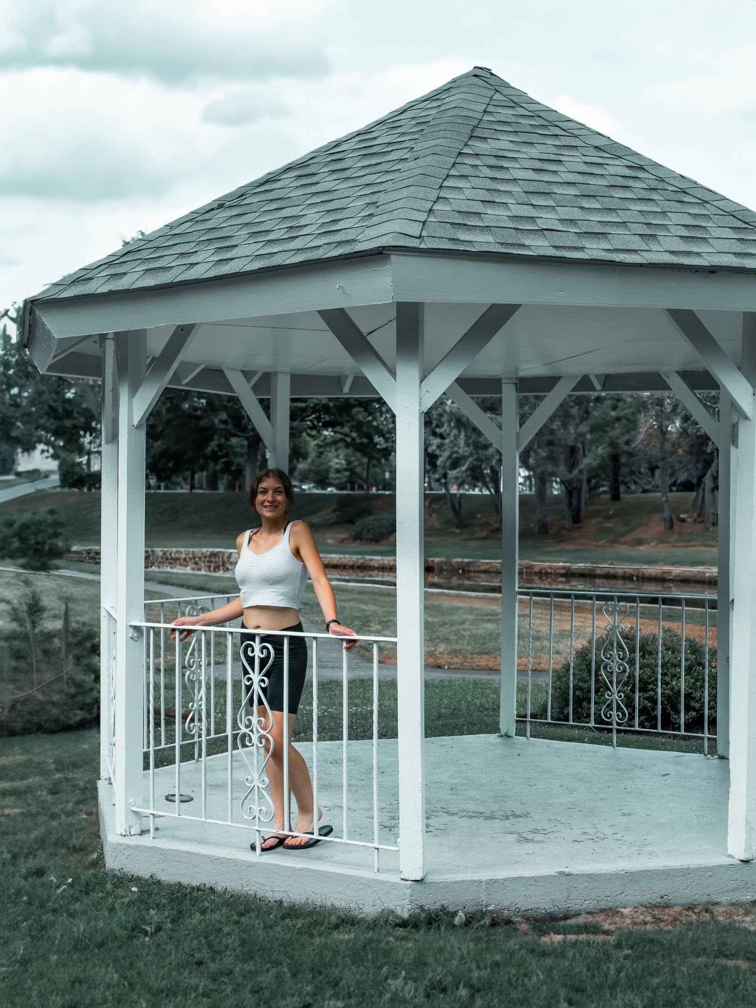 Person posing in a white gazebo with ornate railings on a cloudy day.