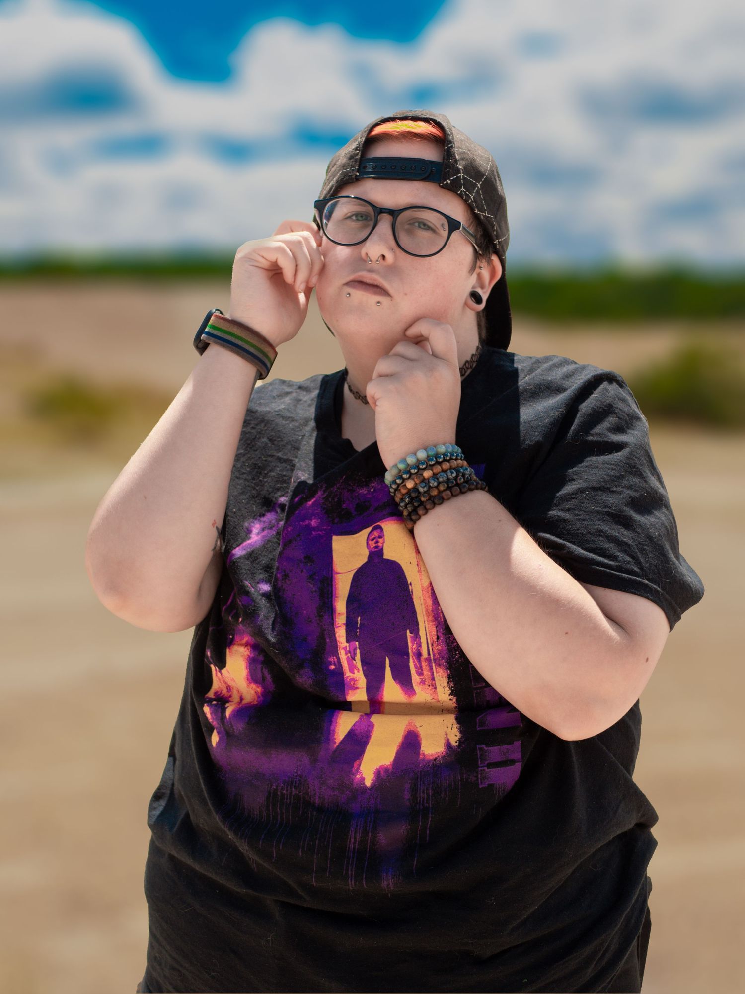 A person wearing glasses and a black t-shirt with a purple graphic design poses outdoors against a cloudy sky.