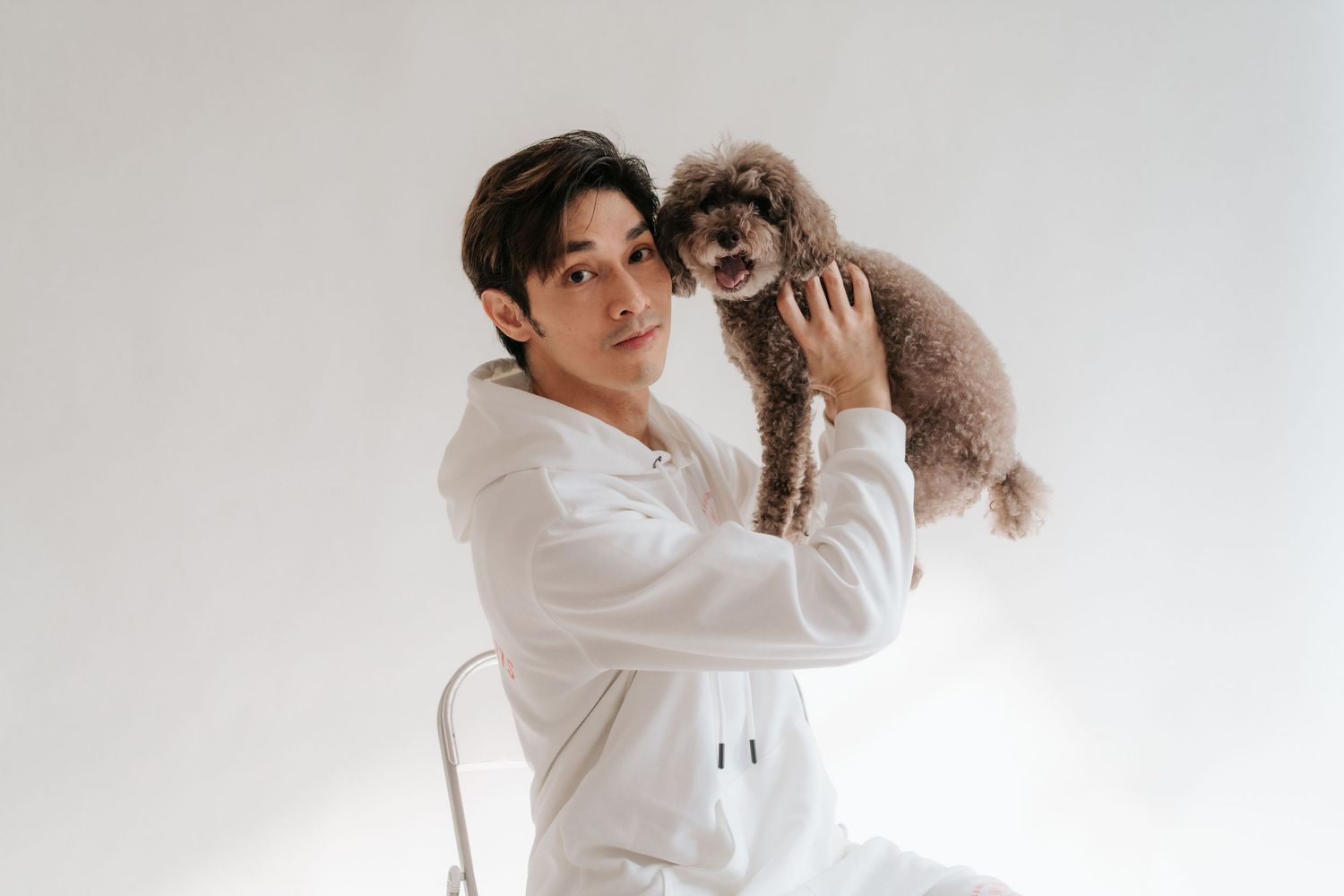 Young Asian man in white hoodie holds a brown toy poodle against a white background.一名身穿白色連帽衫的年輕亞裔男子在白色背景前抱著一隻棕色玩具貴賓犬。
