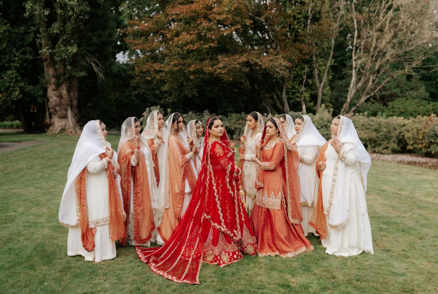A bride in red surrounded by bridesmaids wearing neutral tones on a manicured lawn.