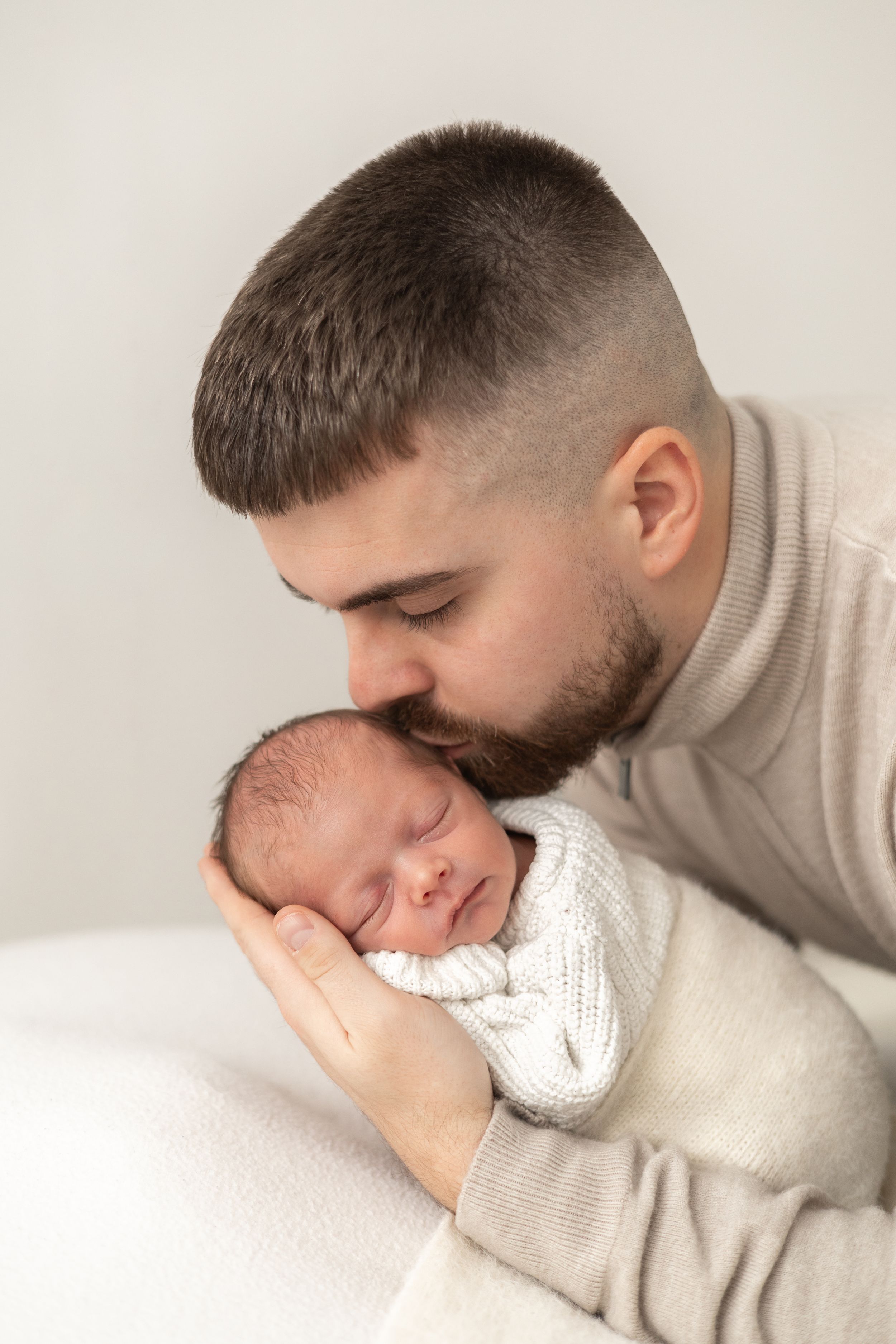Dad gently kisses sleeping newborn baby wrapped in white knit blanket Nottingham baby photographer studio session