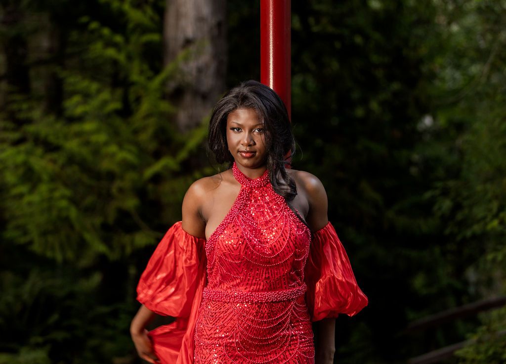 Woman in red halter sequin dress posing confidently beside a red pole in forest.