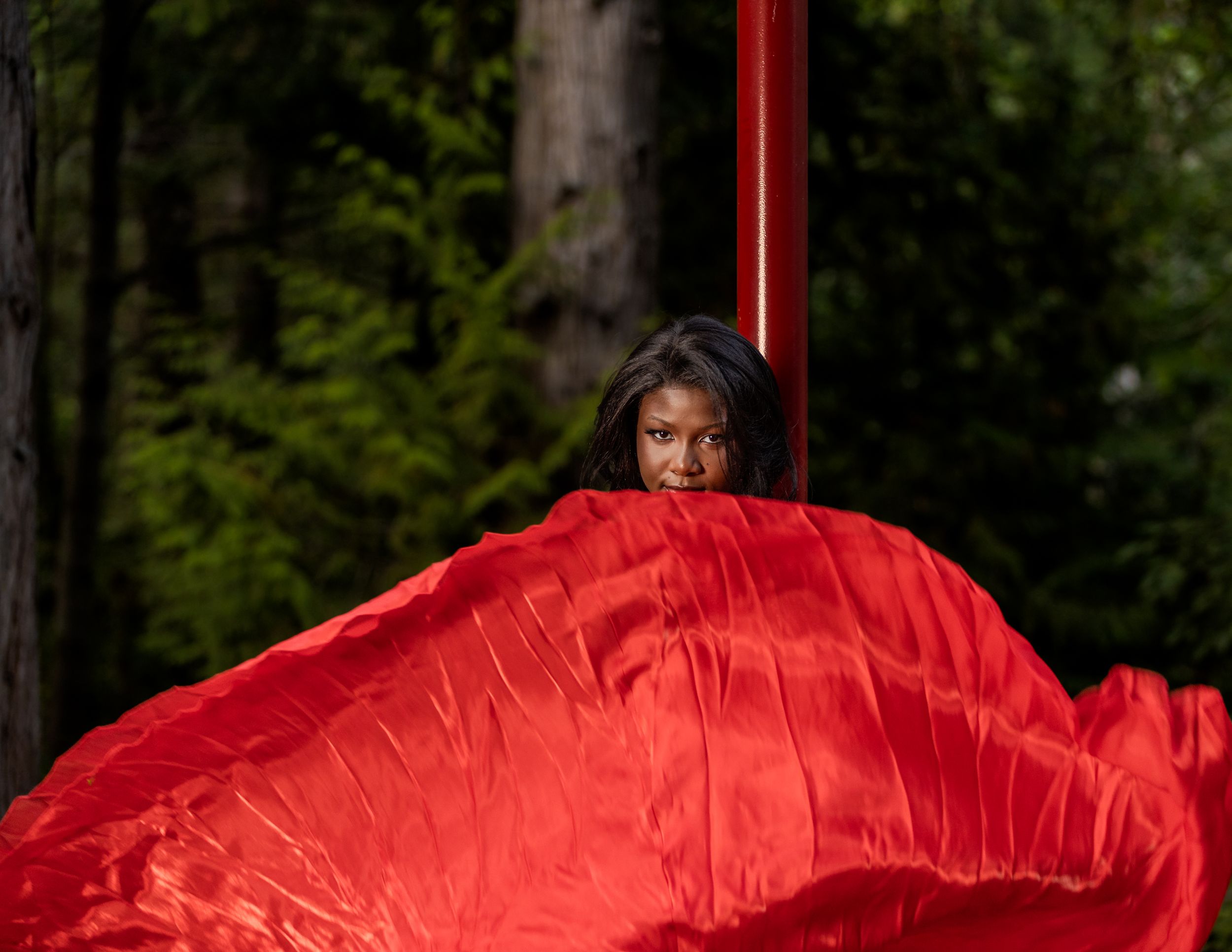 Woman in flowing red fabric poses outdoors among lush green trees, creating a dramatic and elegant visual contrast.