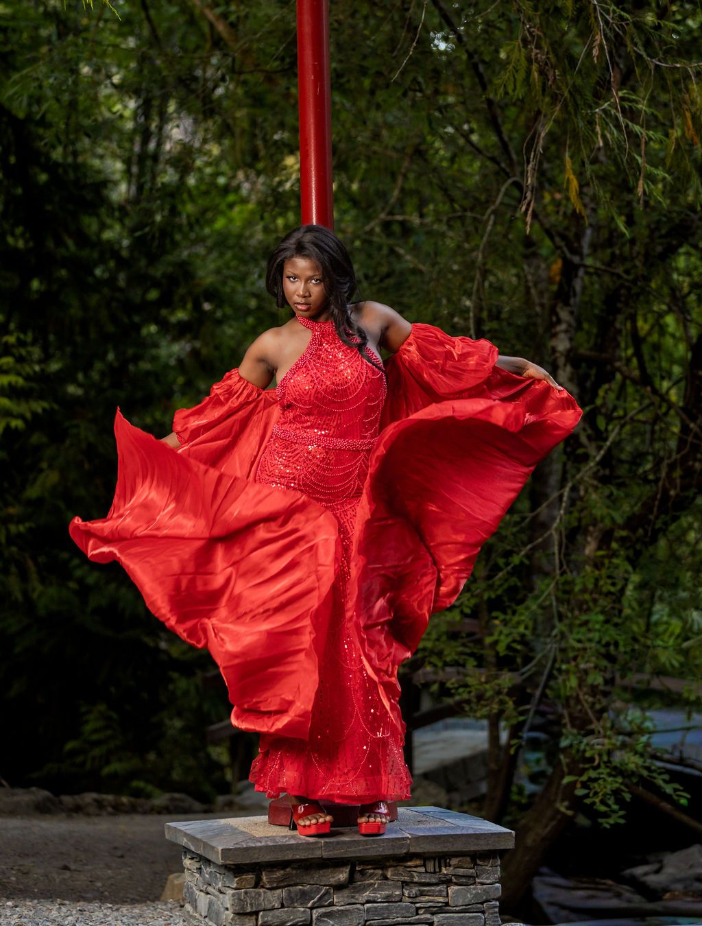 Woman in red sequin gown with flowing fabric billowing in the wind outdoors.
