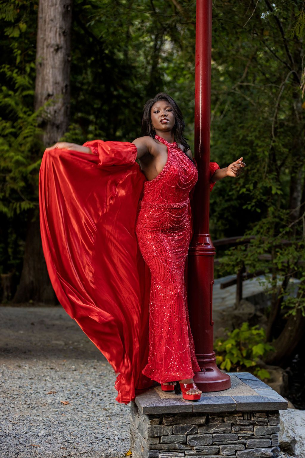 Woman in flowing red sequin gown posing dramatically by a red pole outdoors.