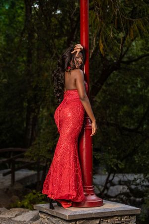 Elegant woman in red sequin mermaid gown posing by red pillar in garden.