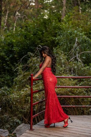 Woman in red sequin backless gown posing on wooden bridge with red railing.