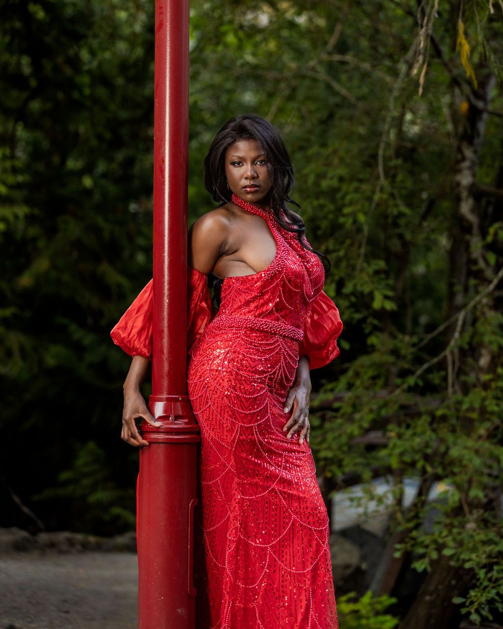 Woman in red beaded gown leaning against a red pole surrounded by green trees.
