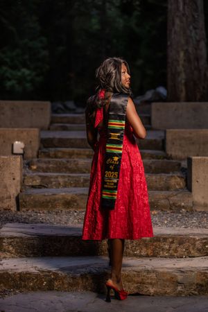 Graduate in red dress with Class of 2025 kente stole standing on stone steps.