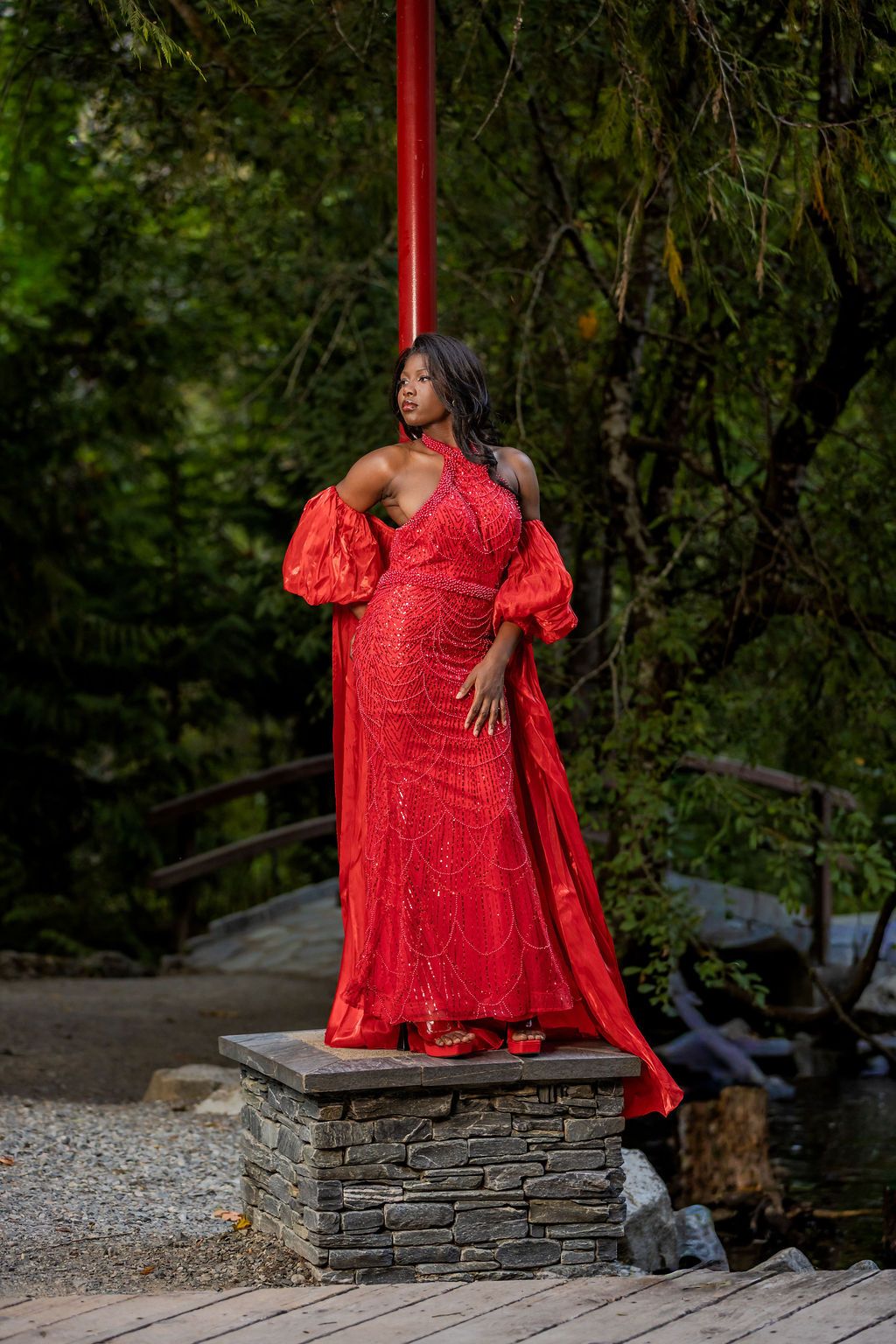 Woman in stunning red sequin gown with puff sleeves standing on stone pedestal in forest.