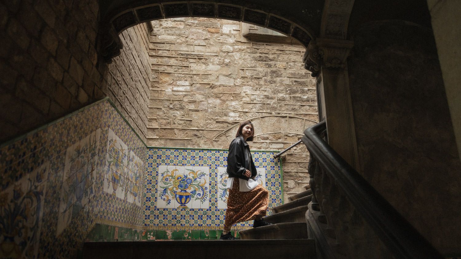 Woman descending ornate tiled staircase in historic building with brick archway and decorative ceramic tile murals.