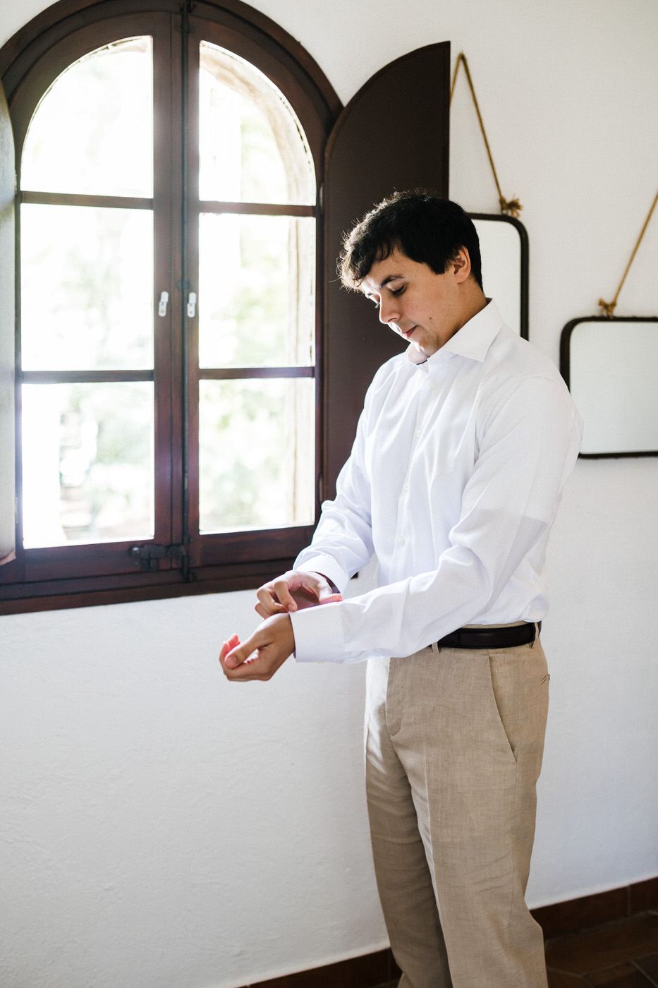 Groom in white shirt and khaki pants adjusts cufflinks by arched window.