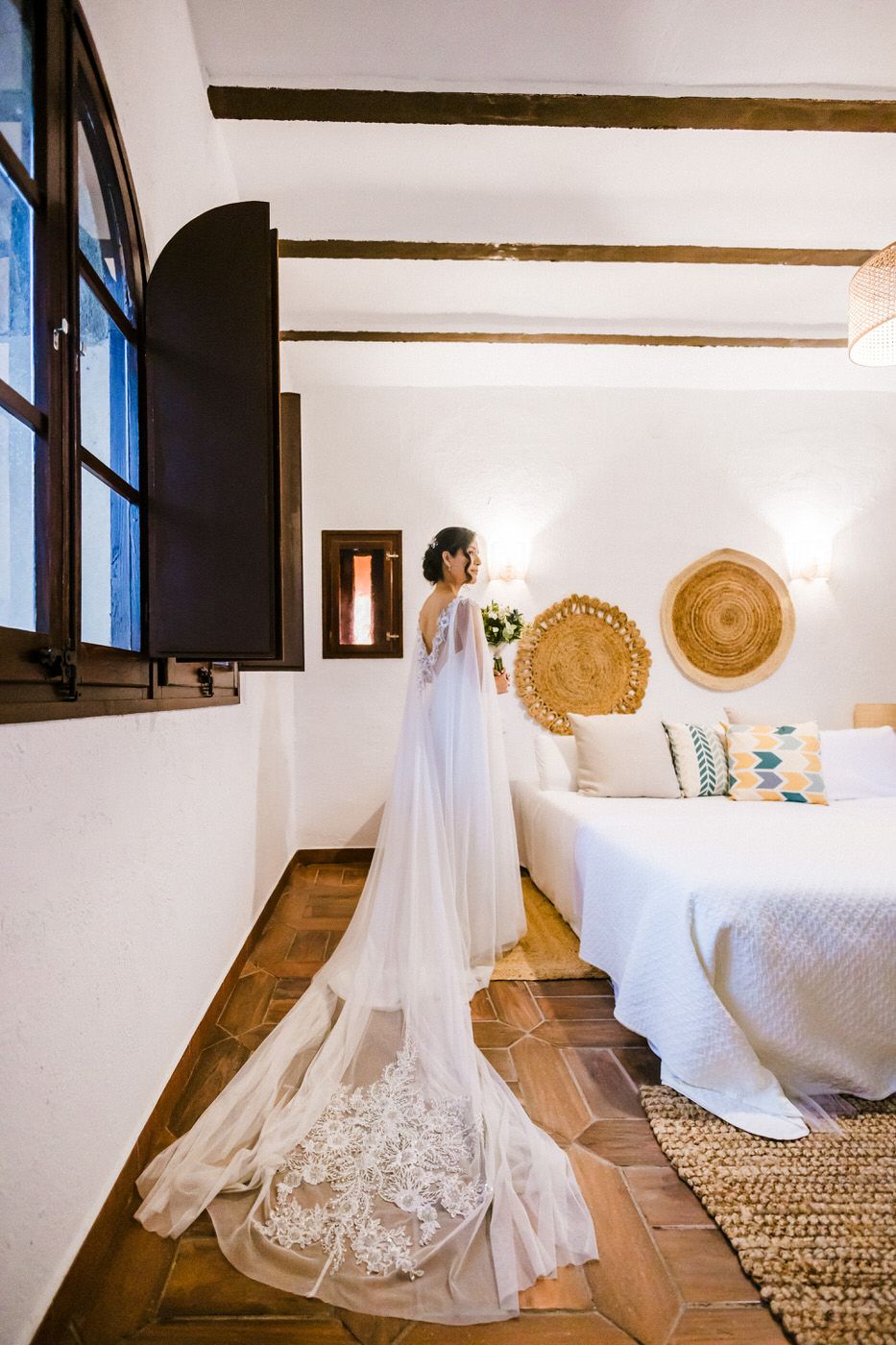 A flowing white wedding dress with delicate lace details displayed on wooden floors in a bright room with natural light.