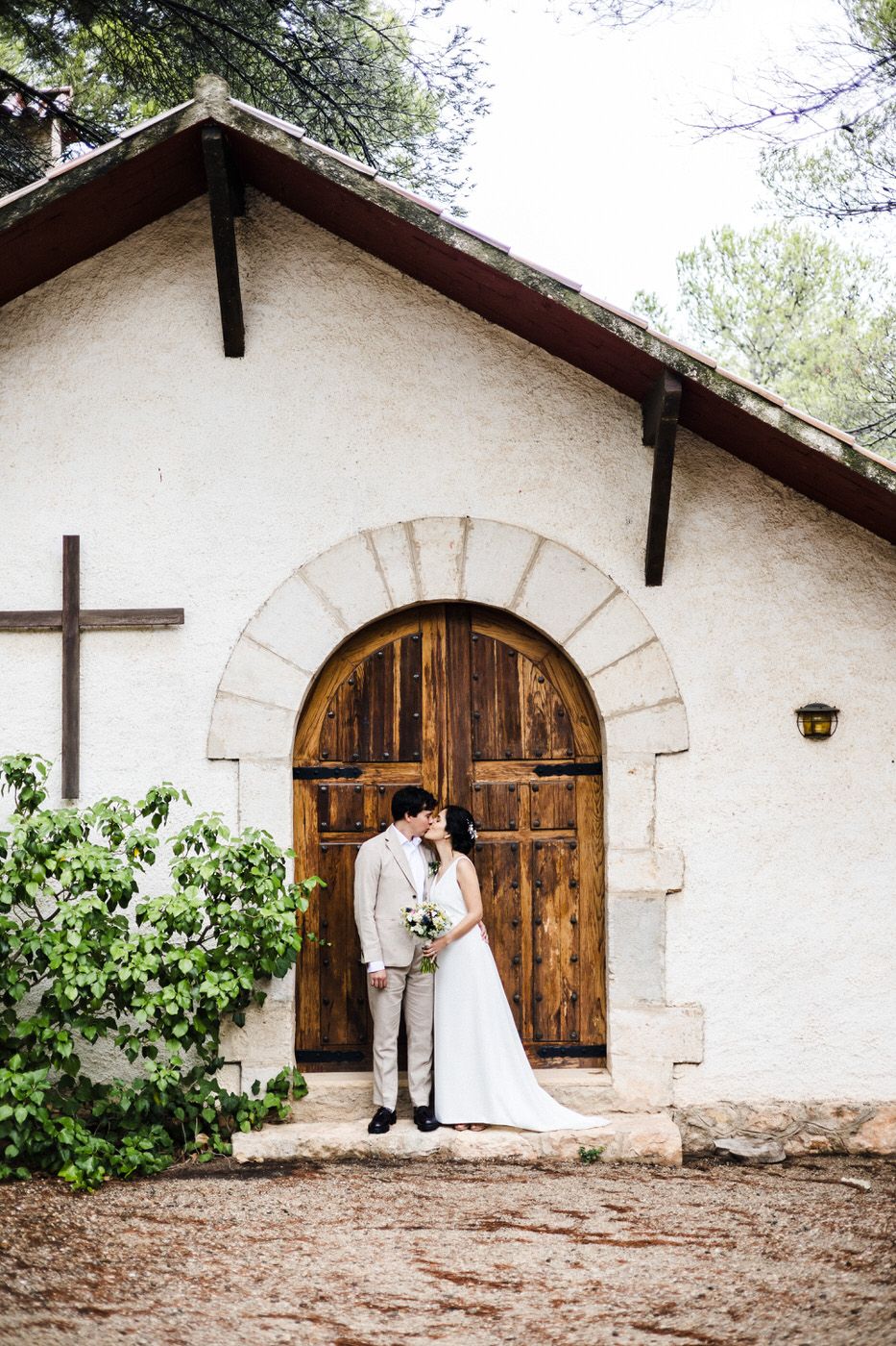 A couple embraces in front of a rustic white church with a wooden arched door and cross on the exterior wall.