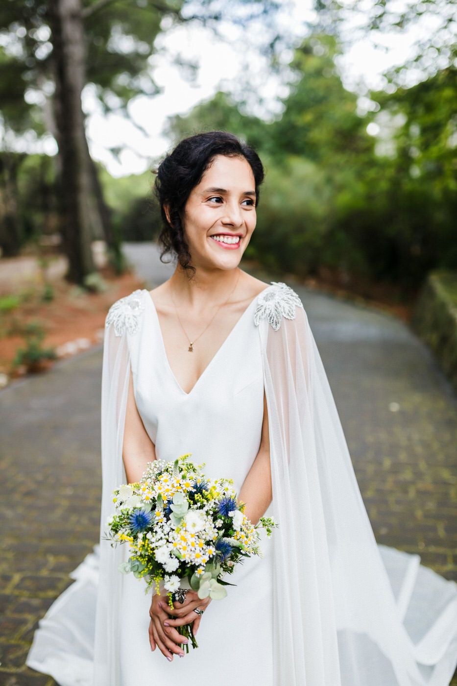 A bride in a flowing white dress holds a yellow and blue bouquet while posing outdoors on a tree-lined path.