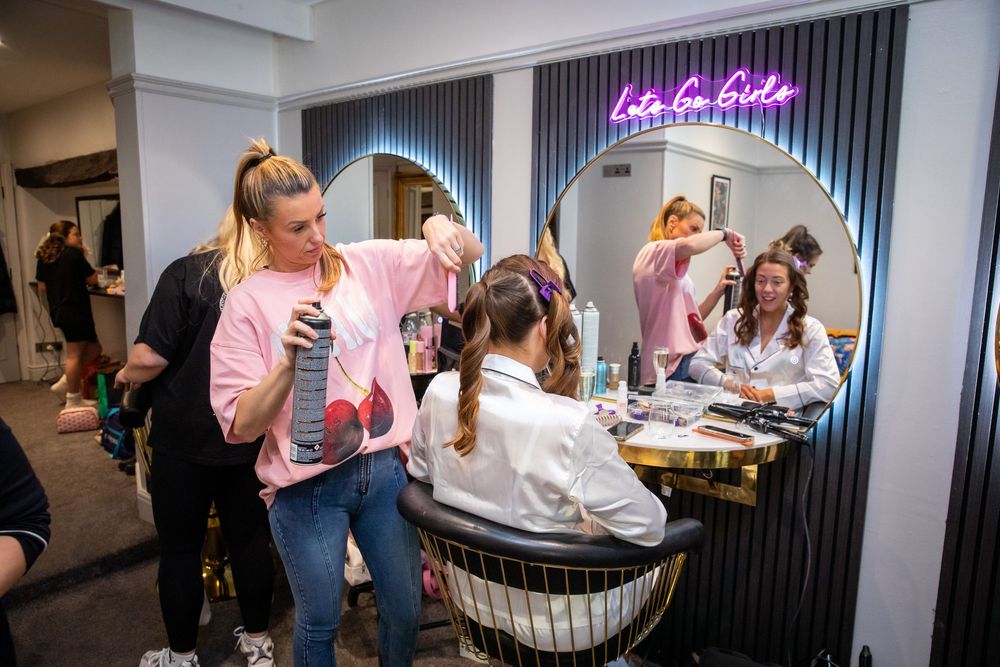 Hair stylist applies hairspray to a seated client in a modern salon with neon sign and illuminated mirrors.