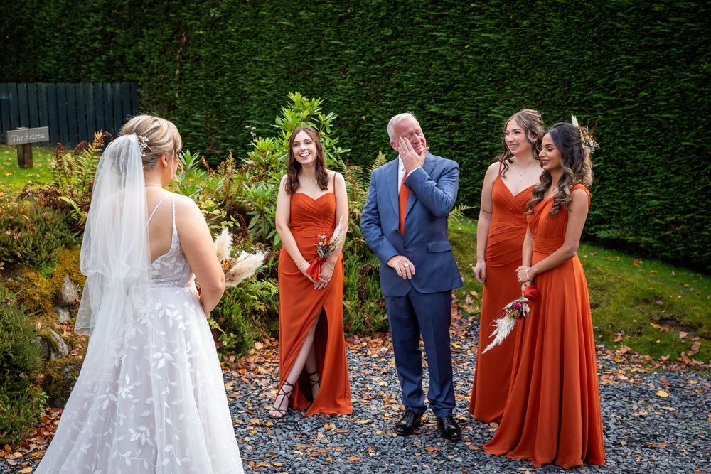 Bride shares emotional first look with bridesmaids in rust orange dresses and father outside on gravel path.