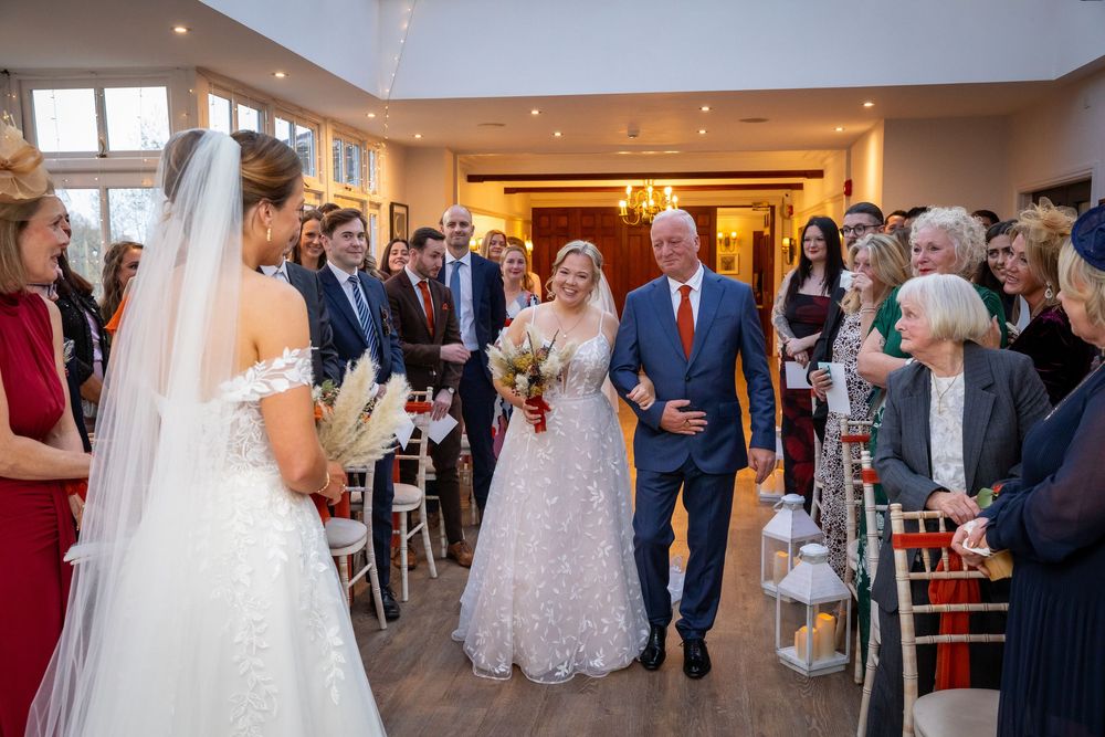 Bride walks down the aisle escorted by father past seated wedding guests in elegant venue.